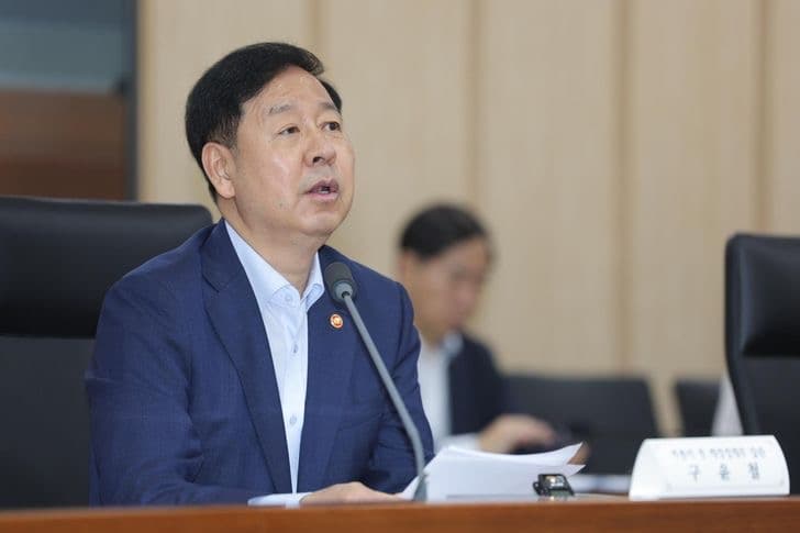 Finance Minister Koo Yun-cheol speaking into a microphone at a government meeting, papers in front of him
