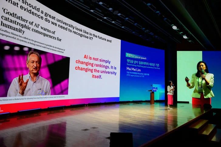 A speaker holds a microphone on stage in front of a large screen during a conference.