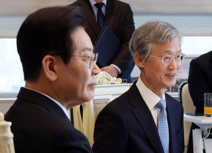 President Lee Jae-myung sits beside a bespectacled man indoors as others stand in the background.