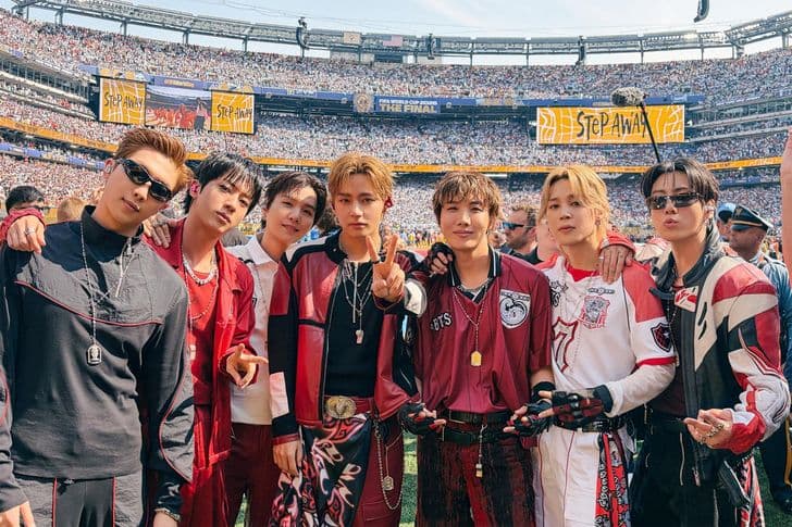 The seven members of BTS pose together on a stadium field before a crowd.