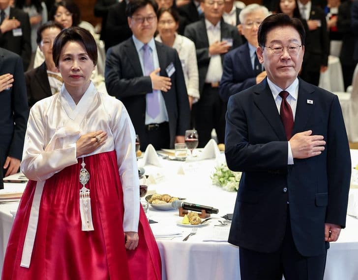 President Lee Jae-myung stands with his spouse, who wears a hanbok, hands over their hearts at an event.