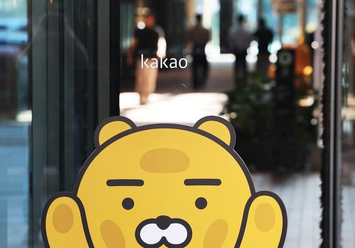 A Ryan character sticker and the kakao logo are seen on a glass storefront door as pedestrians pass in the background.