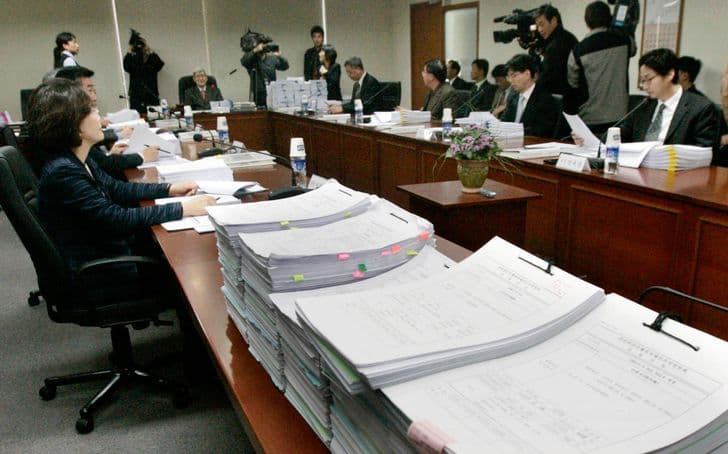 A commission meets around a conference table stacked high with bundles of documents.