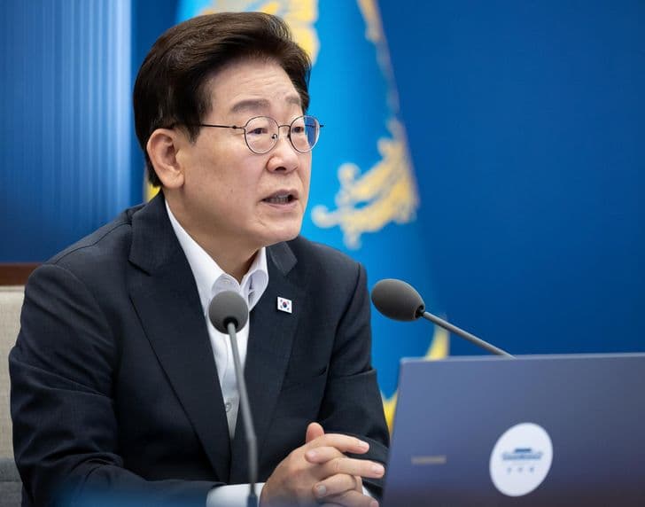 President Lee Jae-myung speaks at a meeting, hands clasped, before the presidential standard.