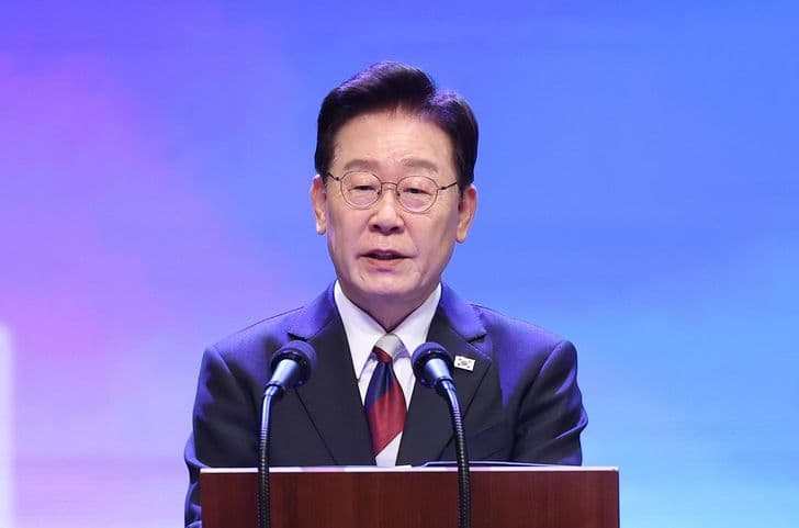 President Lee Jae-myung delivers a speech at a lectern before a blue and purple backdrop.