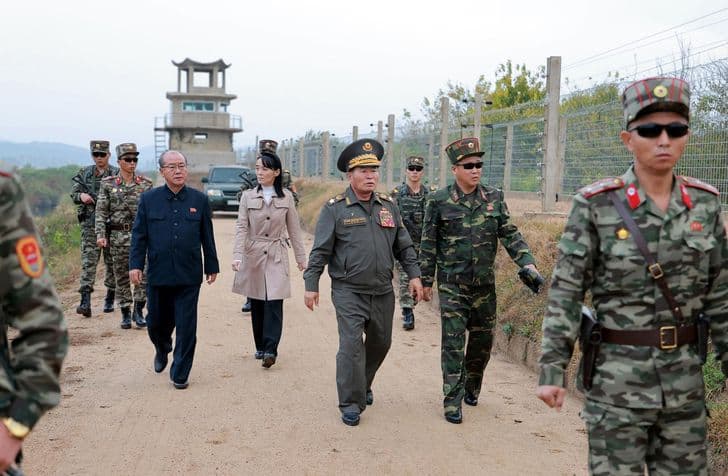 Military officers and officials walk along a fenced border inspection road.