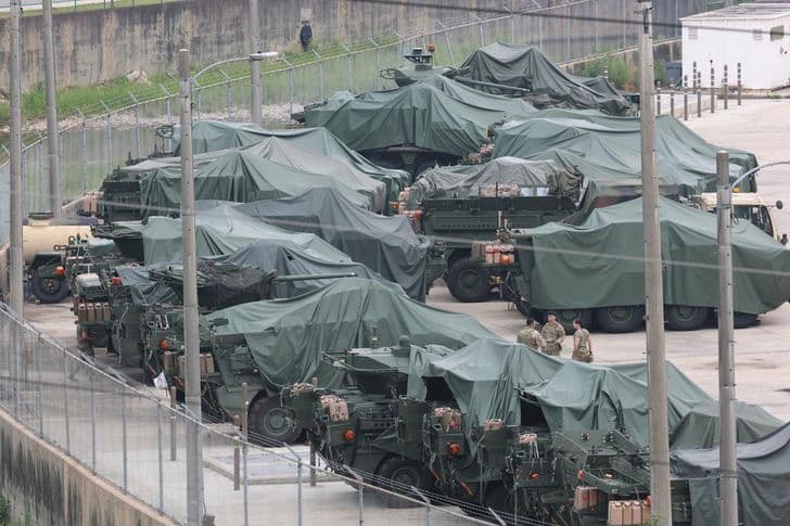 Military vehicles covered in green tarps sit lined up behind a chain-link fence at a base.